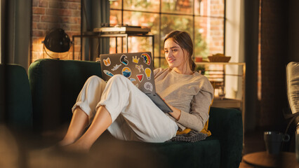 Young Beautiful Woman Lying on Couch, Using Laptop Computer in Stylish Loft Apartment on a Sunny Day. Creative Female Smiling, Checking Social Media, Typing Message. Urban City View from Big Window.