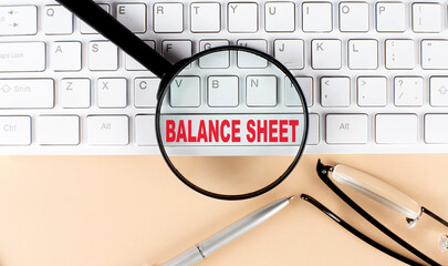Text BALANCE SHEET on keyboard with magnifier , glasses and pen on beige background