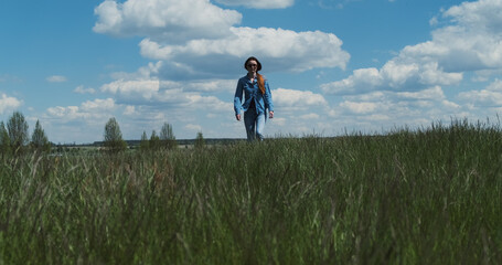 Young woman in denim is walking towards the green tall grass. White clouds, blue sky, spring summer. Concept of return, go to your goal, confidence, countryside. Wide shot, daytime, sunny.