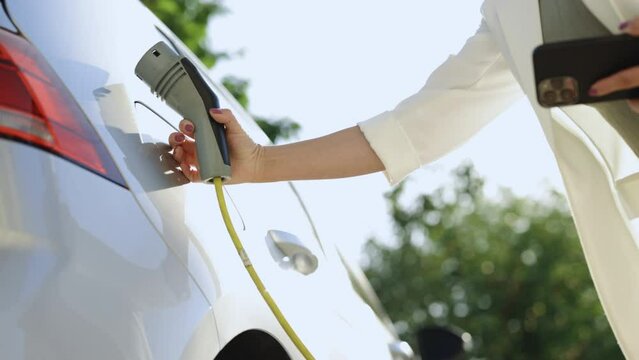 Close up of female hands using smart phone for remote control to charge the modern electric car. Woman's hands unplugging her electric automobile and closing the hatch on charging station