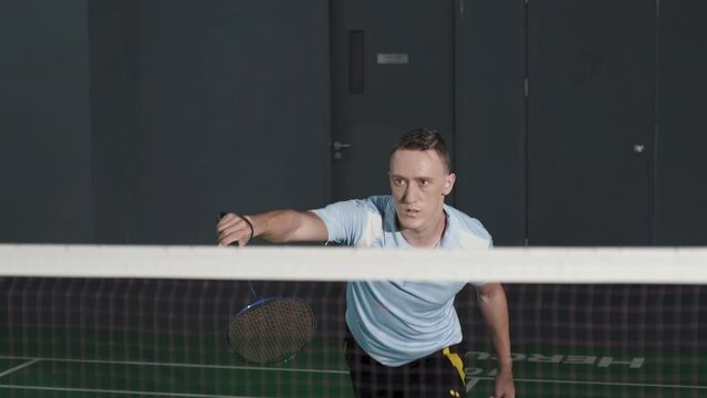 Slow motion shot of Caucasian man in sportswear hitting shuttlecock with racket while playing badminton on indoor tennis court