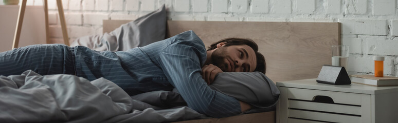 Bearded man looking at alarm clock near water and pills on bedside table in bedroom, banner