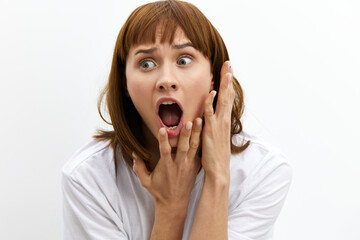 a frightened, shocked, emotional woman screams loudly clutching her face and with very frightened, wide-open eyes stands on a light background in a white T-shirt.