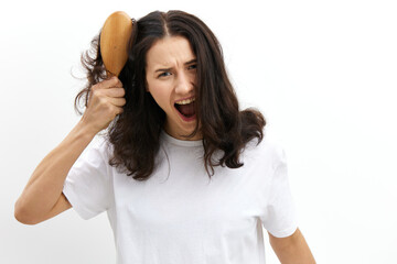 sad, upset woman screams in pain trying to comb her long, black hair with a massage comb, looking at the camera