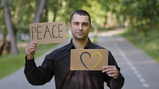 Handsome caucasian man adult businessman patriot guy stands in park outdoors holds two banners painted cardboard with written inscription peace artwork drawing love symbol blue yellow romantic heart