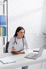 Doctor holding notebook and looking at laptop near clipboard in clinic