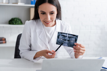 Smiling doctor pointing at scan of teeth near laptop in clinic