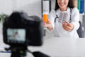 Cropped view of smiling doctor holding pills near blurred digital camera in clinic