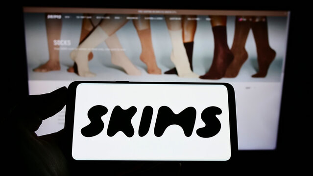 Stuttgart, Germany - 01-28-2022: Person holding mobile phone with logo of American clothing company Skims Body Inc. on screen in front of business web page. Focus on phone display.