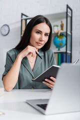 Teacher holding pen and notebook during online lecture on laptop in school