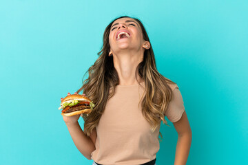 Young woman holding a burger over isolated background laughing