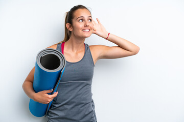 Young sport woman going to yoga classes while holding a mat isolated on white background listening to something by putting hand on the ear