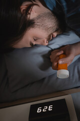 Side view of young man with sleep disorder holding pills near alarm clock in bedroom at night