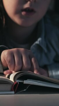 Vertical video. Kid reading hobby. Child education. Imagination development. Closeup of unrecognizable smart little girl studying with open book late at desk.