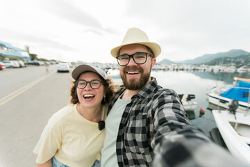 Fototapeta premium Young couple taking a self portrait laughing as they pose close together for camera on their smartphone outdoors in summer port marina with boats and yachts