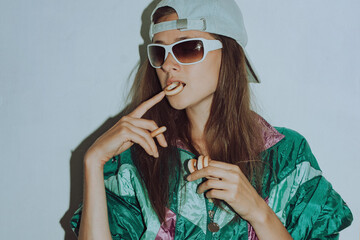 Cool teenager. Fashion girl in colorful trendy jacket and vintage retro sunglasses eating bagels in 80s - 90s style. Teenage girl in a cap on a white background.