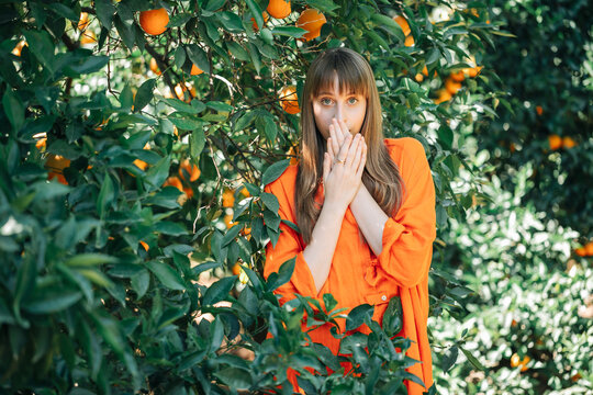 Young Girl In Orange Dress Looking At Camera By Holding Hands In Front Of Mouth In Orange Garden