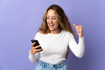 Fototapeta premium Young blonde woman isolated on purple background using mobile phone and doing victory gesture