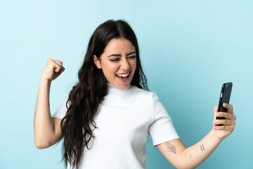 Fototapeta premium Young woman using mobile phone isolated on blue background celebrating a victory