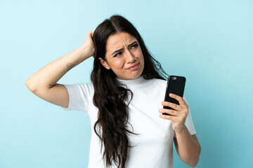 Young woman using mobile phone isolated on blue background having doubts and with confuse face...