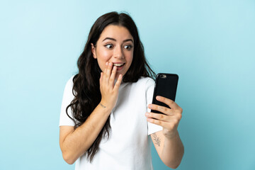 Young woman using mobile phone isolated on blue background with surprise and shocked facial...