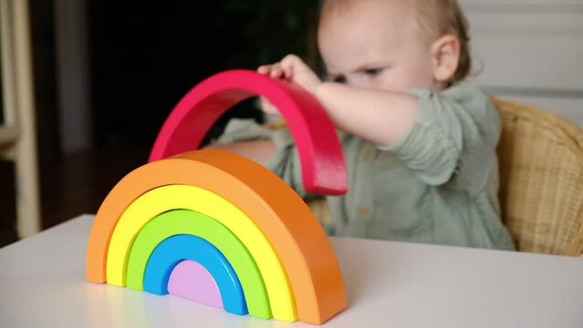 Little baby child in clothes made of natural fabric plays with rainbow colored wooden toys at white table. Cozy room background in scandinavian style. Warm natural colors. Eco friendly lifestyle