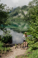 young woman behind the trees looking at the crystal clear water reflection of a lake in the mountains