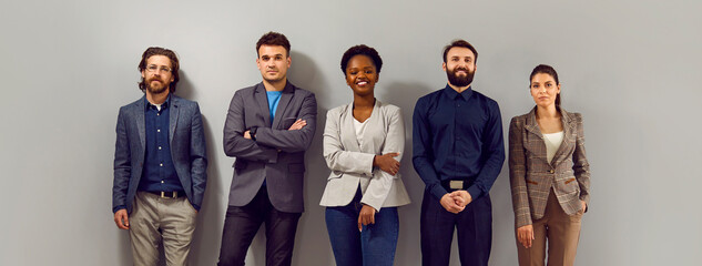 Confident business team in modern smart clothes. Group of happy young diverse people in formal suits standing in fashion studio and leaning on gray wall. Office dress code concept. Banner, header - Powered by Adobe