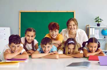 Portrait of cheerful group of elementary school students and their friendly casual female teacher. Joyful schoolchildren leaning on hands laughing at camera with teacher in school classroom.