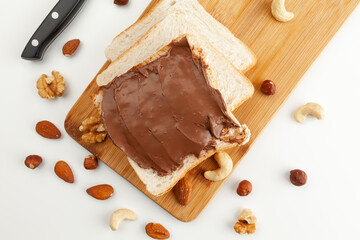 Square bread for toast with chocolate paste on a wooden board. Nuts, a knife and a wooden cutting board with a sandwich and slices of bread on a white table.