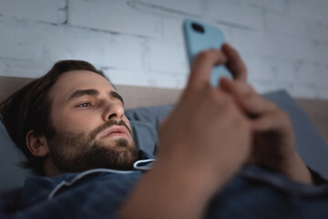 Bearded man in pajama using blurred smartphone on bed at night