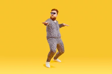Foto op Canvas Plus size male model in funny PJs having fun in modern fashion studio. Happy carefree confident fat bearded man in comfortable leopard pajamas and cool glasses dancing isolated on yellow background © Studio Romantic