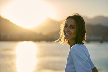 Portrait smiling natural beauty woman looking to camera, enjoying sunset landscape with mountains - 515707780