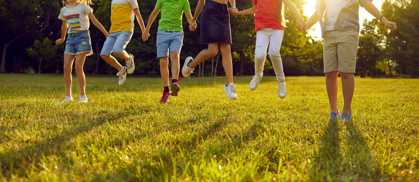 Group of happy boys and girls enjoying summer, playing games and having fun. Several kids holding hands and jumping on fresh green grass in a sunny park. Cropped shot, children's legs. Header, banner