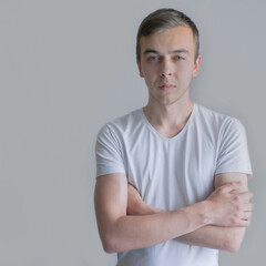 Obraz premium Portrait of a young guy holding crossed arms in a white t-shirt on gray background