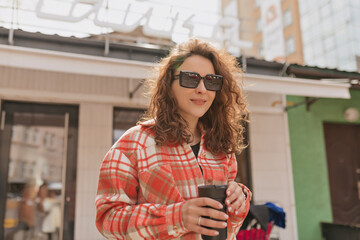 Smiling young caucasian woman walking in morning city in nice warm weather. Brunette wears sunglasses, shirt and jeans. Weekend concept, relaxation, morning with coffee