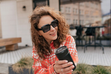 Closeup photo of happy young caucasian woman in sunglasses laughing sitting outdoors. Brunette in good mood wears bright shirt. Lifestyle, different emotions, leisure concept.