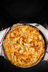 Quiche pie in chef hands on dark background. Savory tart Lorraine with cheese, chicken and mushrooms, freshly baked in oven. Traditional french cuisine. Home cooking recipe. Vertical, copy space