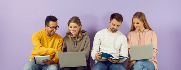 Happy multiracial college or university students sitting in row by purple wall, using laptop...