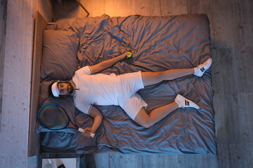 Top view of man in sportswear holding racket and tennis ball while sleeping on bed