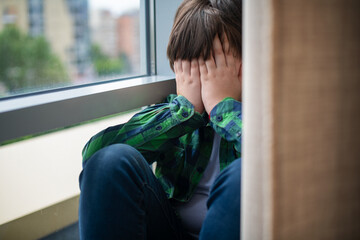 A frustrated, sad and lonely teenager of 12 years old is sitting on the windowsill of the house, covering his face with his hands. Children's social psychology and emotions.