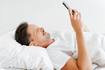 Young smiling man using smartphone, chatting on social media while relaxing on bed. Side view of lazy guy waking up in morning in bedroom and browsing internet on mobile phone