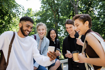 African american student holding smartphone near interracial friends in park.