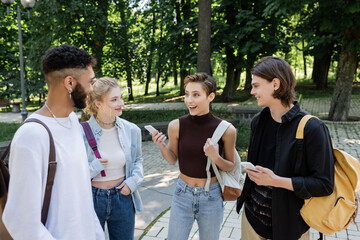 Cheerful student with smartphone talking to interracial friends in park.