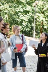 Smiling students with notebooks looking at asian friend with digital tablet in park.
