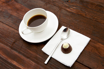 Cap of a coffee, white serviette, chocolate candy and tea spoon on a wooden desk