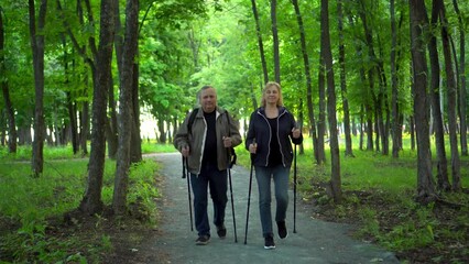 An elderly couple is engaged in Nordic walking in the forest. A man and a woman walk with sticks to improve health. - Powered by Adobe