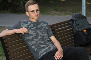 A young man sits on a bench in the shade in summer.