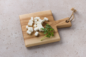 Wooden board with diced feta cheese and a sprig of parsley on a beige textured background. Cooking homemade food