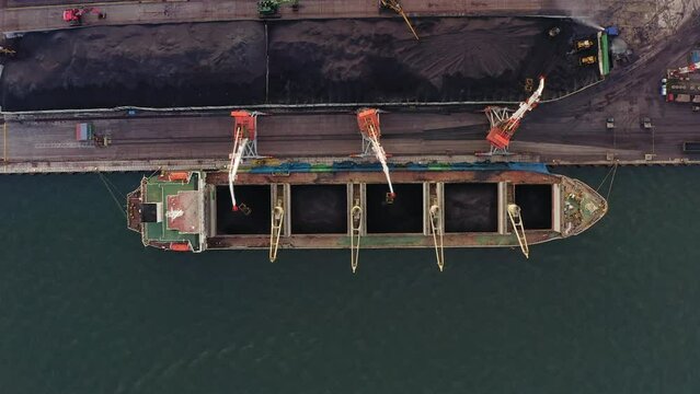 Top view of loading a cargo ship with coal using port cranes. Mooring wall with heaps of coal, evening light. The water gun moistens the coal. Drone view. Time lapse